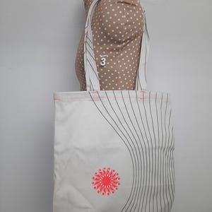 May include: White canvas tote bag with black and pink stripes and a red and pink flower design. The bag has two long straps.
