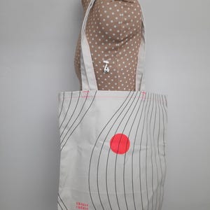May include: White canvas tote bag with black wavy lines and a red circle. The bag has the text "Grazie Cherie" printed on the bottom.