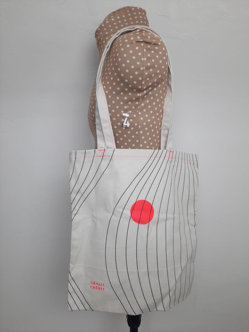 May include: White canvas tote bag with black wavy lines and a red circle. The bag has the text "Grazie Cherie" printed on the bottom.