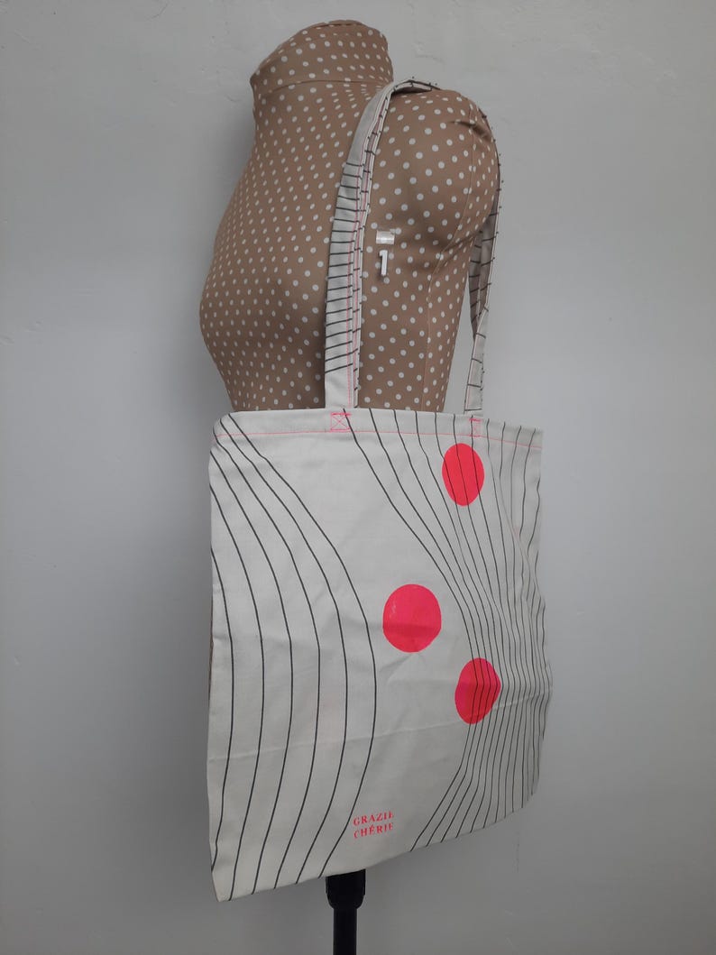 May include: White canvas tote bag with black wavy lines and three bright pink circles. The bag has a pink handle and the text "Grazia Cherry" printed on the bottom.