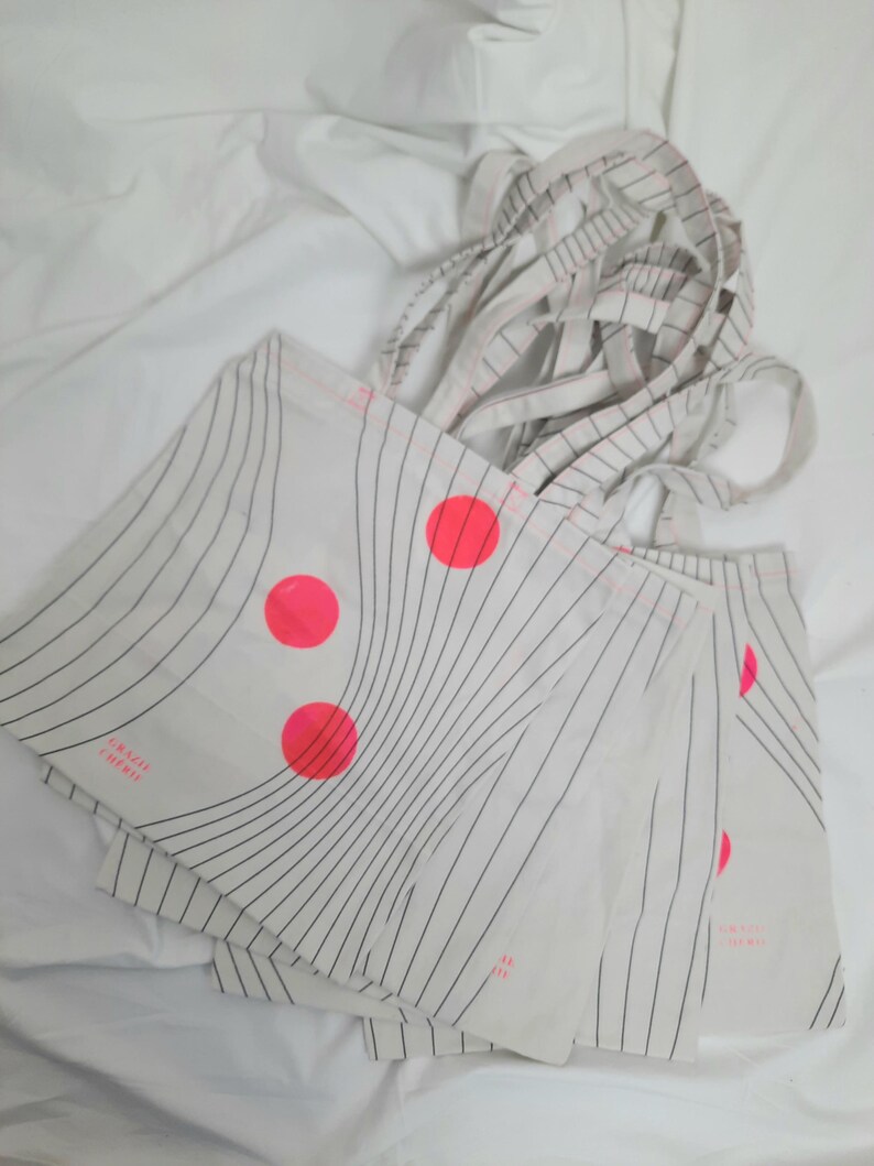 May include: A stack of white tote bags with black stripes and pink circles. The bags have the text "FINISTERRE" printed on them.