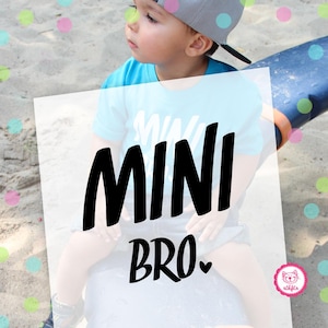 Könnte beinhalten: Ein kleiner Junge trägt ein blaues Shirt mit dem Aufdruck "Mini Bro". Er trägt eine blaue und graue Baseballkappe und sitzt auf einer blauen und grauen Oberfläche.