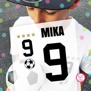 Könnte beinhalten: Weißes Fußballtrikot mit schwarzen Zahlen und dem Namen "Mika" auf der Vorderseite. Das Trikot hat fünf goldene Sterne über der Nummer 9 und fünf goldene Sterne unter der Nummer 9.