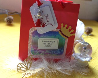 Pode incluir: Uma sacola de presente vermelha com uma etiqueta branca com o nome Julia e um pingente de anjo. Uma etiqueta central diz Kleiner Dankesgruß Ute & David 19.08.2016, decorada com uma coroa amarela, penas brancas, uma borboleta dourada e um pequeno globo de neve.