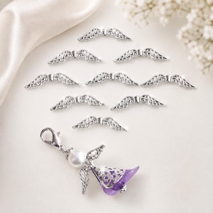 May include: A collection of eight silver-toned metal angel wing charms arranged in a cluster above a single angel-shaped charm. The angel charm features a white spherical bead head, silver wings, and a translucent purple bell-shaped skirt.