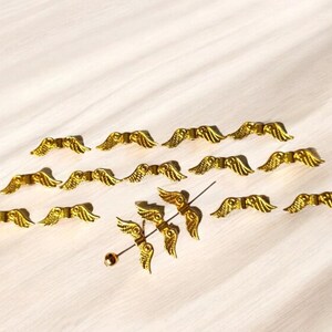 May include: A collection of gold-colored angel wings, some are attached to a pin, others are loose. The wings are a popular craft supply for adding a touch of whimsy to projects.