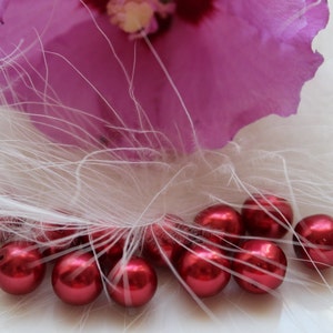 May include: A collection of round, glossy, deep red beads scattered among soft, wispy white feathers. A single large, vibrant purple flower petal with a dark center is positioned behind the feathers, creating a contrast in color and texture.