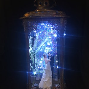 May include: A decorative lantern with a bronze finish houses a wedding cake topper of a couple dancing. The interior is illuminated with blue lights and glitter, creating a romantic ambiance. The base of the topper reads "today".