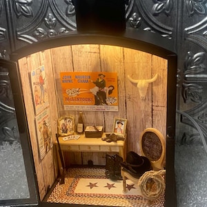 May include: Miniature Western-themed lantern with a detailed interior. Features a desk, chair, cowboy boots, and movie posters. The lantern has a black frame and a handle, with a warm light illuminating the scene.