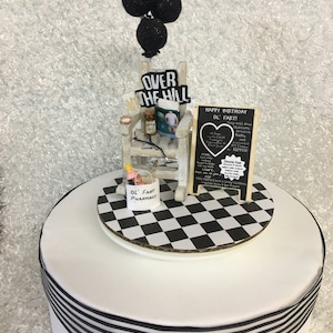 May include: A white cake decorated with a black and white checkered platform, a miniature rocking chair with the words "OVER THE HILL", and a sign that says "HAPPY BIRTHDAY OL' FART!". The cake is topped with black glitter balloons and a pharmacy bag.