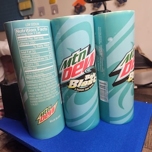 May include: Three blue and green cans of Mountain Dew Baja Blast, a tropical lime flavored soda. The cans have the Mountain Dew logo and the text "Baja Blast" on them. The nutrition facts label is visible on one of the cans.