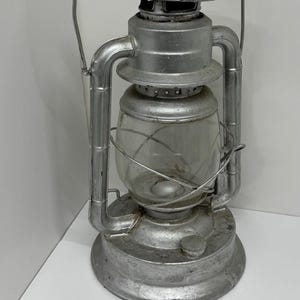 May include: A vintage silver-colored lantern with a clear glass globe. The lantern has a handle for carrying and a small knob at the base. The metal has a slightly weathered appearance, suggesting age and use. The lantern is designed for outdoor use.