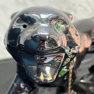 May include: A black panther hood ornament with a green glass eye and a gold chain. The ornament is made of ceramic and has a glossy finish.
