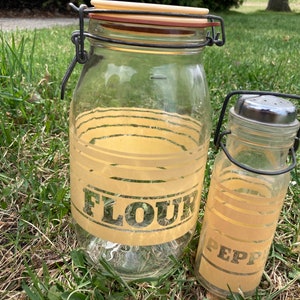 Puede incluir: Dos tarros de vidrio vintage con tapas de metal y asas de alambre. El tarro más grande está etiquetado como "FLOUR" y el más pequeño como "PEPPER". Ambos tarros tienen un diseño a rayas amarillo y blanco.