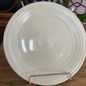 Puede incluir: Un plato de cerámica blanco con un borde elevado y un diseño sutil de círculos concéntricos. El plato está sobre un soporte de madera.