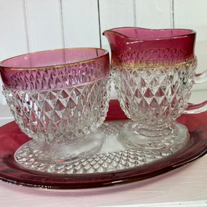 Puede incluir: Un juego vintage de azucarero y jarra de crema de vidrio con un patrón de diamante y una base de color rojo rubí. El juego está en una bandeja de color rojo rubí a juego.