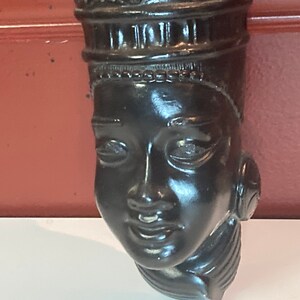 Peut inclure: Sculpture en bois noir représentant la tête d'une femme avec une couronne et des détails complexes. La sculpture a une expression sereine et est une œuvre d'art traditionnelle.