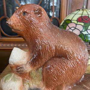 Puede incluir: Una figurita de cerámica de un castor marrón sentado en una base con un hongo blanco. El castor está sosteniendo el hongo en sus patas.