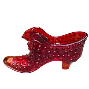 Może przedstawiać: Czerwony, szklany przedmiot dekoracyjny w kształcie buta. But ma teksturowaną powierzchnię z małymi, wypukłymi wypustkami i małym obcasem. Szkło jest w głębokim czerwonym kolorze, na białym tle.