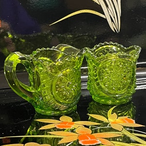 Könnte beinhalten: Zwei grüne Glaskännchen für Sahne und Zucker mit geschliffenem Glasdesign. Die Kännchen stehen auf einer schwarzen Oberfläche mit einem Blumenmuster.