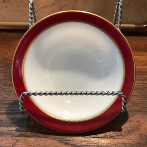 Può includere: Una piccola ciotola in ceramica bianca con un bordo rosso e oro. La ciotola è su un supporto in metallo.