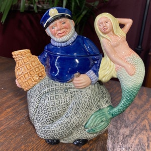 May include: A ceramic teapot shaped like a fisherman holding a basket and a mermaid with a green tail. The fisherman is wearing a blue jacket and a hat. The mermaid is wearing a pink shell bra.