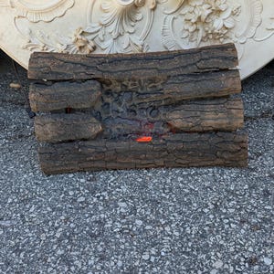 May include: A rectangular artificial fireplace log set featuring a dark brown, textured bark finish. The logs are arranged in a stacked pattern with a central recessed area containing a small, glowing orange ember effect at the bottom centre.
