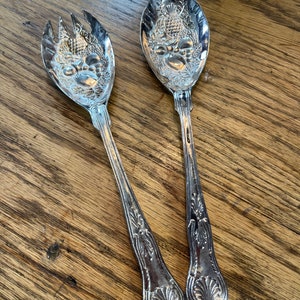 May include: A set of two silver salad serving utensils with ornate handles and a floral design. The utensils are on a wooden surface.