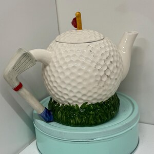 May include: A novelty teapot shaped like a golf ball, with a golf club handle and a spout. The white teapot sits on a green base, atop a light blue tin. A red and yellow flag decorates the lid.