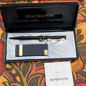 Peut inclure: Un coffret noir et or pour briquet et stylo avec une inscription dorée "REMINGTON". Le coffret est ouvert et le briquet et le stylo sont visibles. Le briquet est un briquet noir et or avec une bordure dorée. Le stylo est un stylo noir et or avec un clip doré. Le coffret est posé sur une surface à motifs.