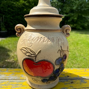 May include: A beige ceramic jar with a lid, featuring a hand-painted design of an apple and grapes. The jar has the word "Ramstorf" written on the front.