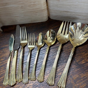 Pode incluir: Um conjunto de oito peças de talheres dourados, incluindo uma faca de manteiga, uma faca de jantar, dois garfos, duas colheres, um garfo de servir e uma colher de servir.