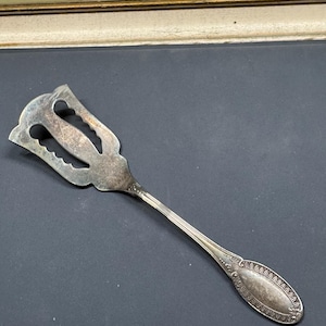 Op de afbeelding: Een vintage zilverkleurig serveerbestek met een decoratieve handgreep en een gleuf, sierlijke kop. De kop heeft een gestileerd ontwerp met uitgesneden vormen. De handgreep heeft een ovaal uiteinde met een gedetailleerd patroon. Het bestek wordt waarschijnlijk gebruikt om voedsel te serveren.