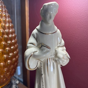Peut inclure: Figurine en céramique crème représentant une figure religieuse tenant un livre et une colombe. La figurine a des bordures et des détails dorés sur la robe et les bords. La figure se tient devant un mur bordeaux.