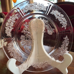 Peut inclure: Une assiette en verre rose avec un motif floral en relief de roses. L'assiette est sur un support blanc.