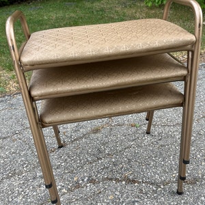 Puede incluir: Un taburete de metal de tres niveles con acabado dorado y tapicería de tela marrón con estampado. El taburete tiene un diseño sencillo con una parte superior rectangular y tres patas.