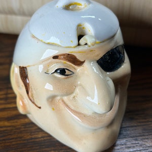May include: A ceramic sugar or creamer dispenser shaped like a pirate's head with a white hat and an eyepatch. The pirate has a brown mustache and a mischievous smile.