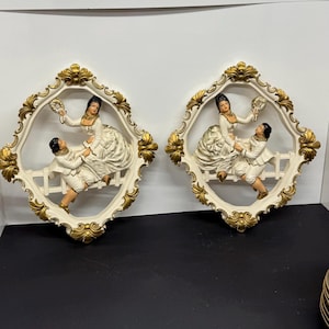 May include: A pair of decorative wall plaques. Each plaque features a cream-coloured couple in period dress, set within a white frame with gold detailing. The figures are posed on a small white fence.