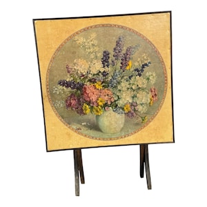 May include: A vintage floral print on a square wooden tray with a black frame. The print features a bouquet of colorful flowers in a white vase, set against a neutral background. The tray is supported by a wooden stand.