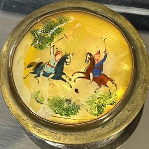 Puede incluir: Una caja redonda de latón con una escena pintada de dos personas a caballo cabalgando por un paisaje. La caja está decorada con un borde dorado y tiene una tapa de vidrio transparente.