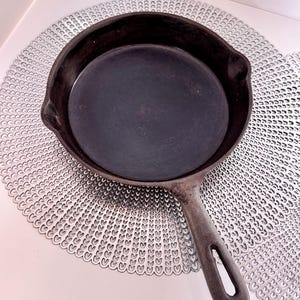 May include: A black cast iron skillet with a single handle. The skillet is sitting on a white surface with a decorative pattern.