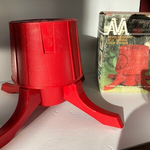 Può includere: Un supporto per albero di Natale in plastica rossa con una base a quattro punte. Il supporto è etichettato "Multi-Use Stand for Christmas and All Year" e "AVA Revolutionary New Spring Lock System".