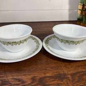 Könnte beinhalten: Zwei weiße Teetassen mit grünem Blumendesign am Rand, jede auf einer passenden Untertasse mit dem gleichen Muster.