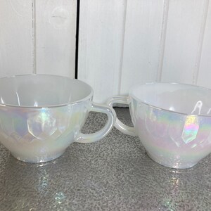 May include: Two white iridescent teacups with a geometric pattern. The cups have a handle and are made of glass.