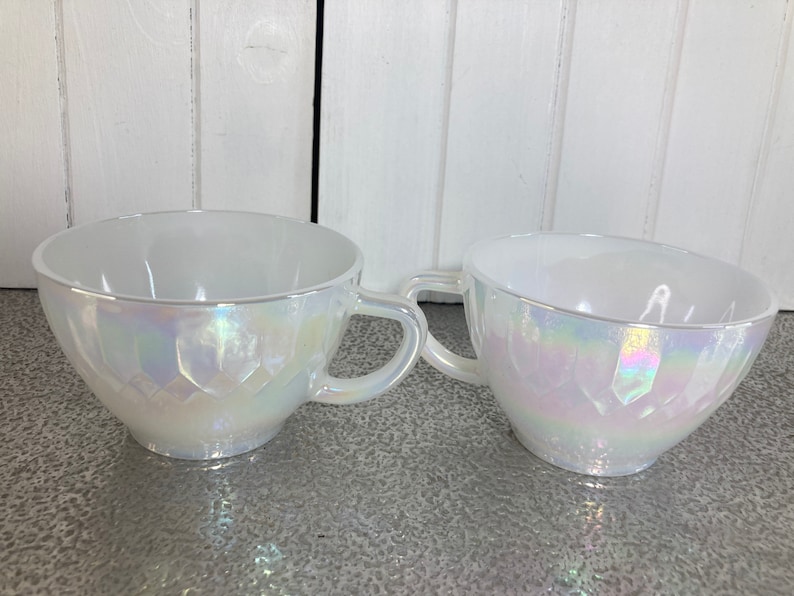 May include: Two white iridescent teacups with a geometric pattern. The cups have a handle and are made of glass.