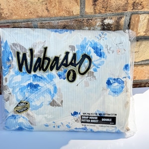 May include: A packaged double fitted sheet with a floral pattern in shades of blue and gray on a white background. The brand name "Wabasso" is displayed in black script. The packaging includes text indicating the sheet is made in Canada.