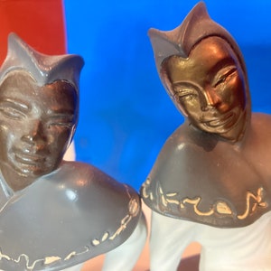 May include: Two ceramic figurines with bronze-colored faces and gray collars with gold accents. The figures are dressed in white and have a stylized, smiling expression. The background is a mix of red and blue.