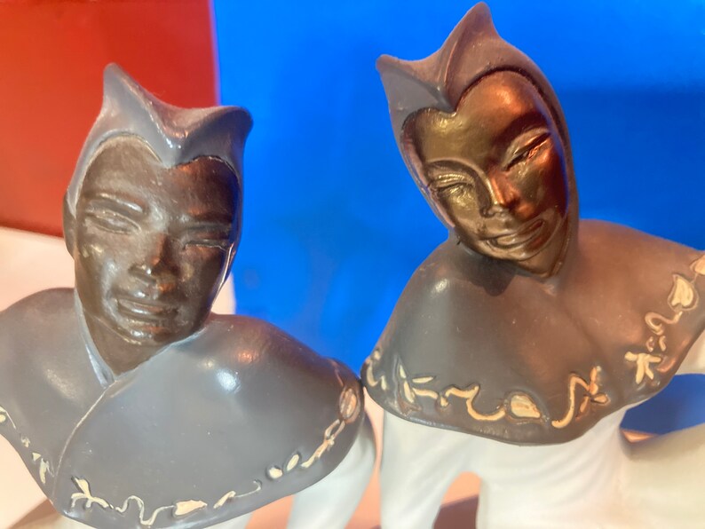 May include: Two ceramic figurines with bronze-colored faces and gray collars with gold accents. The figures are dressed in white and have a stylized, smiling expression. The background is a mix of red and blue.