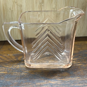 May include: A clear glass pitcher with a square shape and a textured design of overlapping arrows. The pitcher has a handle and a spout.
