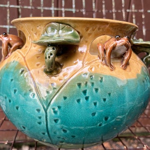 May include: A ceramic planter with a green and brown glaze. The planter is decorated with three frogs and a mushroom. The planter is perfect for adding a touch of whimsy to your home.